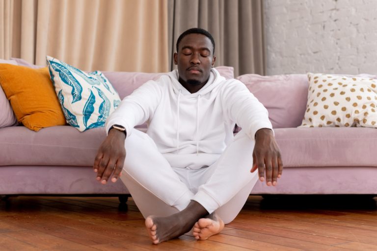 Calm African American male in white clothes sitting with closed eyes and crossed legs on floor against comfortable sofa in living room meditating